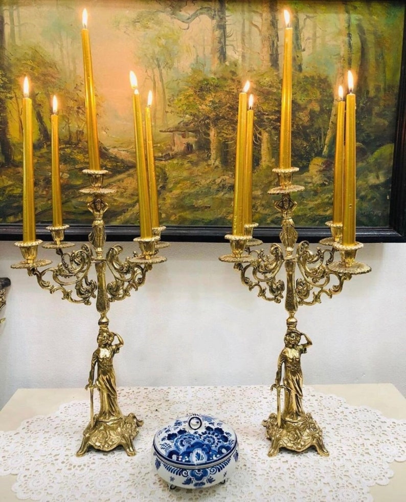 May include: A pair of gold metal candle holders with ornate designs and a female figure on each base. Each holder has six candle arms and is decorated with intricate scrollwork. The candle holders are displayed on a white lace tablecloth with a blue and white porcelain trinket box in the center.