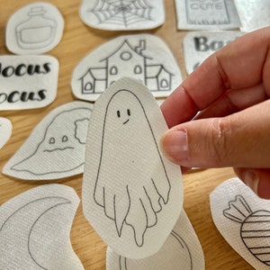 May include: A white ghost with a smiling face and a simple outline is drawn on a white paper sticker. The sticker is being held by a hand.