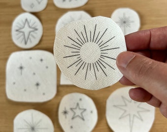 May include: A set of nine white paper circles with black ink drawings of stars and a sunburst design. The circles are arranged in a semi-circle on a wooden surface.