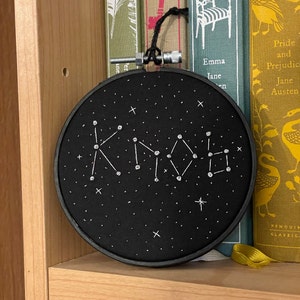 May include: A black embroidery hoop with a constellation design and the word "Kindred" stitched in white thread. The background is a starry night pattern. The hoop is displayed in front of a bookshelf with classic book titles.