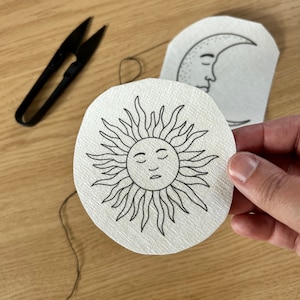 May include: A circular sun design with a face and radiating lines, and a crescent moon design, both printed on white fabric. Black scissors and thread are also visible. The designs are likely for embroidery or crafting.