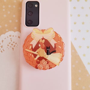 May include: A pink phone case with a pop socket featuring a cartoon witch riding a broom with a black cat. The pop socket is decorated with a wreath of leaves and a yellow and pink bow.