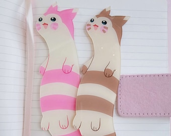 May include: Two long, thin, plastic bookmarks shaped like a cartoon cat with pink and brown striped tails. The bookmarks are on a pink and white lined notebook.