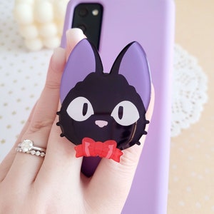 May include: A black cat phone grip with a red bow tie. The phone grip is attached to a purple phone case.