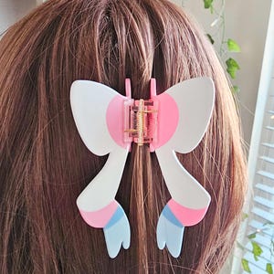May include: A white and pink bow-shaped hair clip with blue accents. The clip is in the shape of a bow with two long tails that are curled at the ends. The bow is white with pink accents and the tails are blue with pink accents.