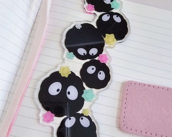May include: A clear acrylic bookmark with five black and sparkly round characters with white eyes. The characters are surrounded by colorful glitter stars.