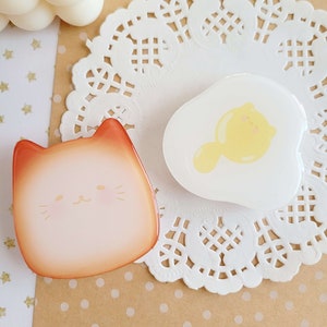 May include: Two resin cat-shaped charms. One charm is orange and white with a cartoon cat face. The other charm is white with a yellow cartoon cat inside a white egg shape.