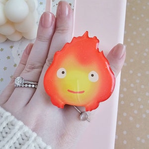 May include: A pink phone case with a red and yellow fire spirit phone grip. The fire spirit has a smiling face and is made of resin.