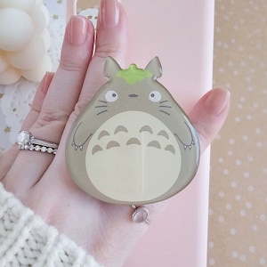 May include: A light pink phone case with a gray and green Totoro phone grip attached to the back.