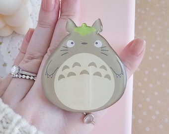 以下が含まれることがあります： 薄いピンクの携帯電話ケースに、グレーとグリーンのトトロの携帯電話グリップが背面に装着されています