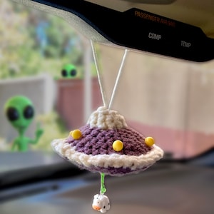 May include: A purple and white crocheted UFO car charm with yellow accents hanging from a rearview mirror. A small white crocheted cow is attached to the bottom of the UFO.