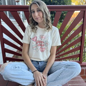 Könnte beinhalten: Ein cremefarbenes T-Shirt mit den Worten "You Are" und den Worten "Enough, Loved, Capable, Worthy, Giving" in Herzformen. Das Shirt wird mit hellblauen Jeans kombiniert.