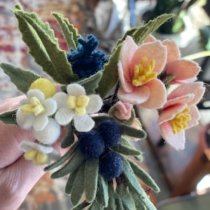 May include: A felt flower bouquet with pink, white, yellow, and blue flowers. The bouquet also includes green leaves and dark blue berries.