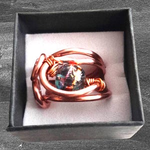 May include: A copper-colored wire-wrapped ring with a central iridescent glass bead. The ring is presented in a black gift box with a white interior, showcasing the handcrafted jewelry.