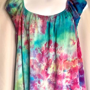 May include: A short-sleeved blouse featuring a vibrant tie-dye pattern in shades of bright pink, turquoise, purple, and lime green. The garment has a wide, rounded scoop neckline with gathered fabric details along the collar and short cap sleeves.
