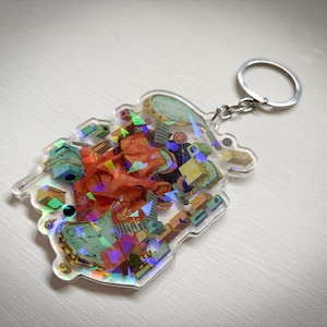 May include: A clear keychain with a colourful, abstract design featuring a red, orange and yellow figure. The keychain is made of acrylic and has a silver metal ring.