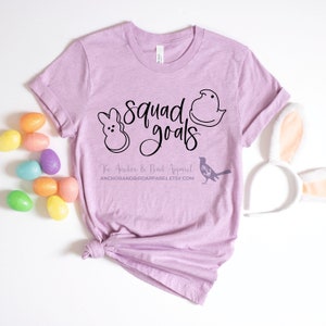 Peut inclure: Un t-shirt violet clair avec le texte "squad goals" en écriture noire. Le t-shirt présente deux contours noirs de bonbons en guimauve Peeps. Le texte "The Anchor & Bird Apparel" et une adresse de site Web sont imprimés sous le graphique.