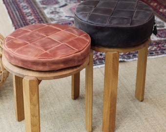 May include: Two wooden stools with round seats, each topped with a circular, quilted leather cushion. One cushion is a warm reddish-brown, while the other is a dark charcoal gray. The stools feature simple, sturdy legs and a smooth finish.