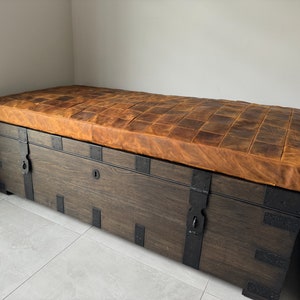May include: A dark brown wooden storage bench with a brown leather top. The bench has metal accents and hinges.