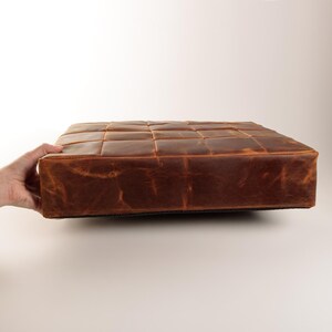 May include: A square, brown leather cushion featuring a grid-like stitched pattern on the top surface. The leather has a distressed, matte finish with visible natural grain variations and tonal shading across the entire surface area.