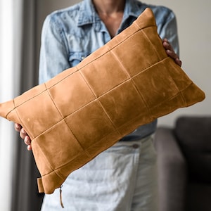 May include: A rectangular tan leather pillow cover featuring a grid-like patchwork design with visible stitching. The cover has a smooth, matte texture and includes a small leather zipper pull tab at the bottom corner for closure and access.