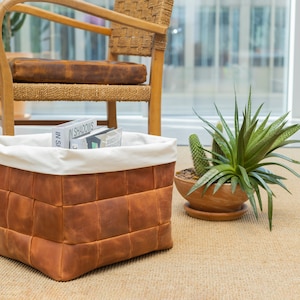 May include: A brown leather storage basket with a white fabric lining. The basket is filled with books and is sitting on a light brown rug. There are two potted plants in the background.