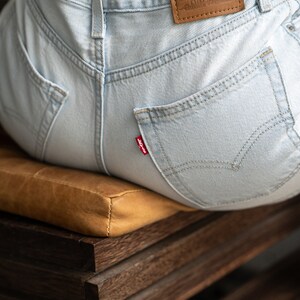 May include: Light blue denim jeans featuring a tan leather patch on the back waistband with the text Levi Strauss & Co. Original Riveted Genuine Quality Clothing. A small red tab with the word Levi's is attached to the right back pocket.