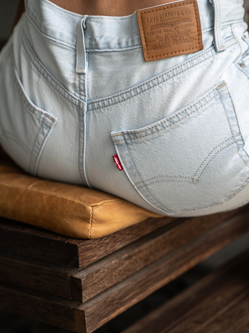 May include: Light blue denim jeans featuring a tan leather patch on the back waistband with the text Levi Strauss & Co. Original Riveted Genuine Quality Clothing. A small red tab with the word Levi's is attached to the right back pocket.