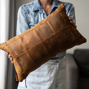 May include: A brown leather pillow with a quilted design. The pillow is made of multiple squares of leather sewn together.