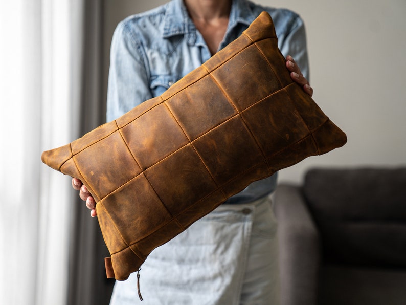 May include: A brown leather pillow with a quilted design. The pillow is made of multiple squares of leather sewn together.