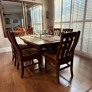 K&ouml;nnte beinhalten: Ein Esstisch aus Holz mit sechs St&uuml;hlen. Der Tisch hat eine dunkle Holzoptik und die St&uuml;hle eine helle Holzoptik. Der Tisch ist mit einem Platzdeckchen und einer Glassch&uuml;ssel gedeckt. Der Raum hat einen Hartholzboden und wei&szlig;e Fensterl&auml;den.