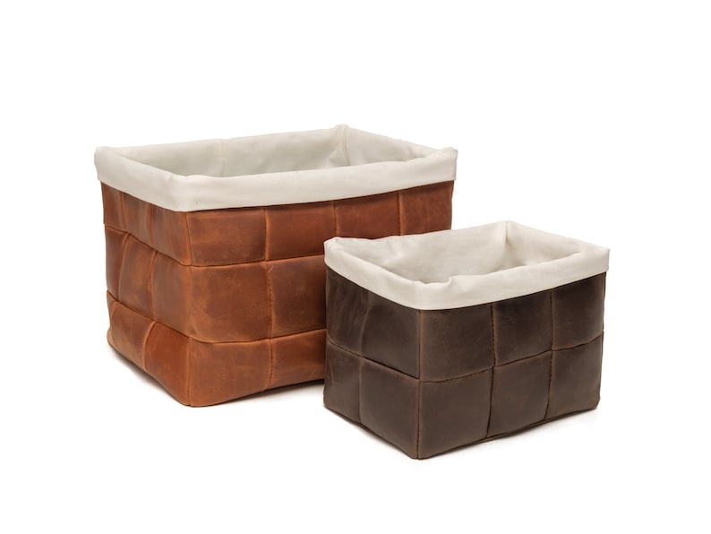 May include: Two woven leather storage baskets with white fabric lining. The larger basket is brown and the smaller basket is dark brown.