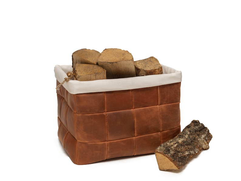 May include: A brown leather woven storage basket with a white fabric liner, filled with pieces of firewood. A single piece of firewood sits in front of the basket.