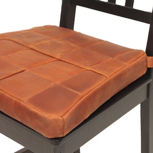 K&ouml;nnte beinhalten: Ein braunes Leder-Sitzkissen mit gestepptem Design. Das Kissen ist mit einem Riemen an einem schwarzen Holzstuhl befestigt.