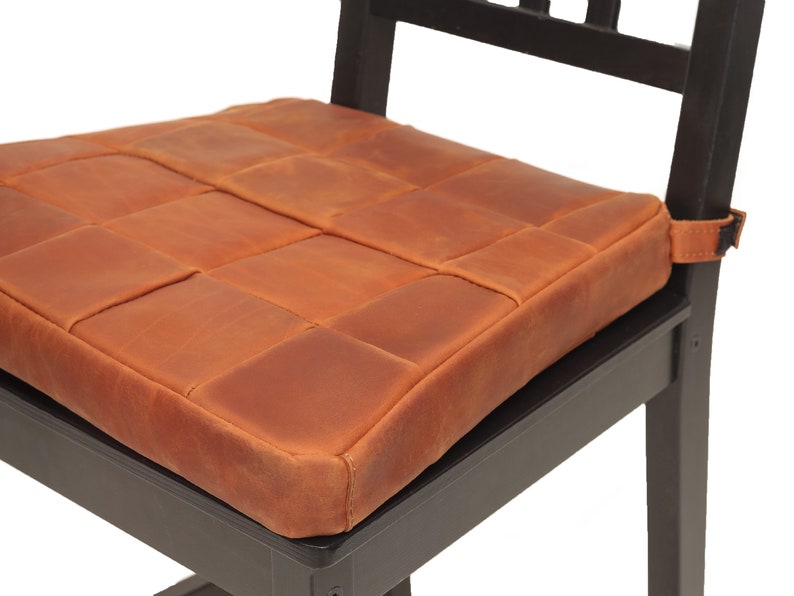 K&ouml;nnte beinhalten: Ein braunes Leder-Sitzkissen mit gestepptem Design. Das Kissen ist mit einem Riemen an einem schwarzen Holzstuhl befestigt.