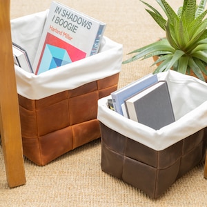 May include: Two brown leather storage baskets with white fabric liners. The basket on the left contains books, including one titled "In Shadows I Boogie" by Hartland Miller. The basket on the right contains a stack of books.