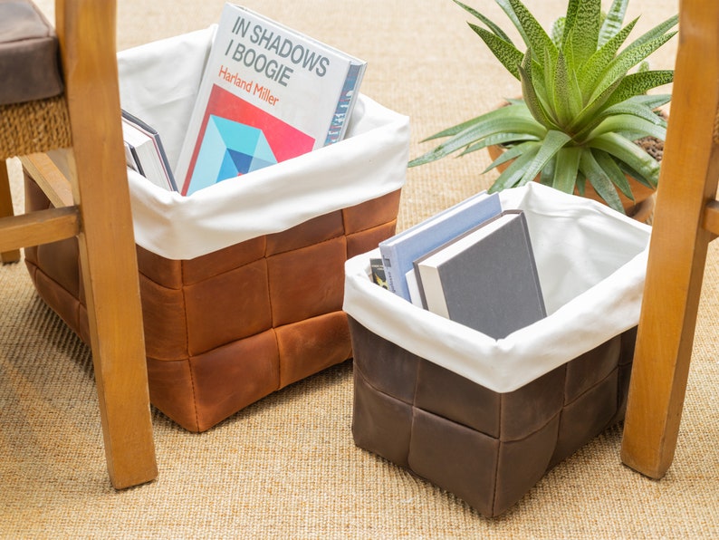 May include: Two brown leather storage baskets with white fabric liners. The basket on the left contains books, including one titled "In Shadows I Boogie" by Hartland Miller. The basket on the right contains a stack of books.