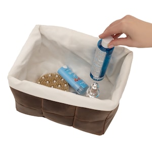 May include: A brown faux leather storage basket with a white fabric lining. The basket is filled with various toiletries, including a blue bottle, a white tube, a brown brush, and a silver bottle.