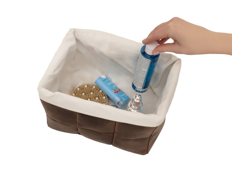 May include: A brown faux leather storage basket with a white fabric lining. The basket is filled with various toiletries, including a blue bottle, a white tube, a brown brush, and a silver bottle.