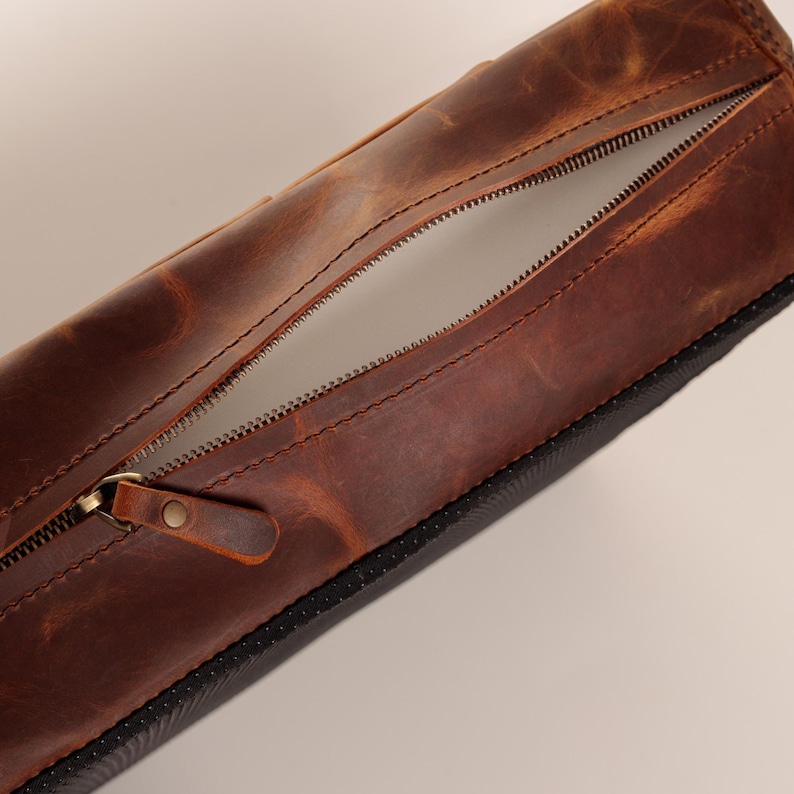 May include: A cylindrical brown leather pencil case featuring a long metal zip closure with a matching leather pull tab. The leather has a distressed, variegated texture in shades of deep brown and tan, with a black textured base.