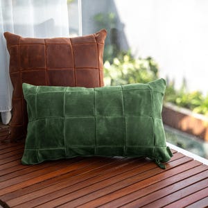May include: A square brown leather pillow and a rectangular green suede pillow, both featuring a grid-like stitched pattern. The brown pillow is positioned behind the green pillow, and both items have visible zippers on their lower right corners.
