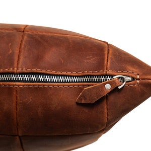 May include: A brown leather pillow with a zipper closure and a leather pull tab. The pillow is made of multiple pieces of leather sewn together.