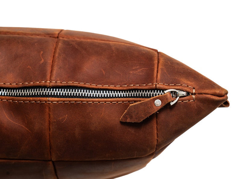 May include: A brown leather pillow with a zipper closure and a leather pull tab. The pillow is made of multiple pieces of leather sewn together.