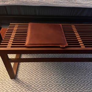 May include: A wooden bench with a brown leather cushion. The bench is sitting on a white and gray patterned rug.
