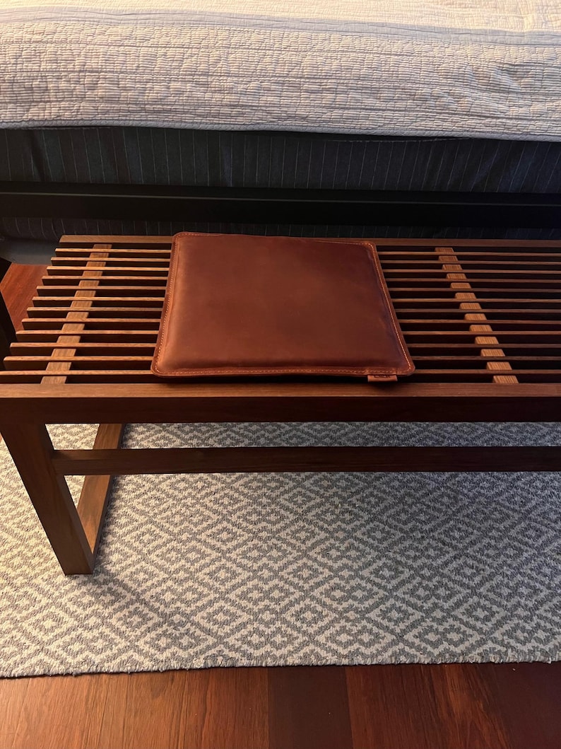 May include: A wooden bench with a brown leather cushion. The bench is sitting on a white and gray patterned rug.