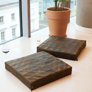 May include: Two square, brown leather cushions with a woven pattern. A glass of red wine and a small glass of amber liquid sit nearby. A potted plant in a terracotta pot is in the background.