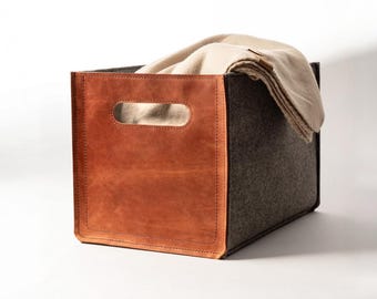 May include: A rectangular storage bin featuring dark grey felt sides and a smooth tan leather front panel with a cutout handle. A folded beige fabric blanket is draped over the back edge of the bin, partially filling the interior space.