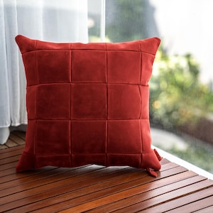 May include: A square, brick-red decorative pillow with a grid pattern. The pillow is made of a soft, velvety material and is placed on a wooden surface. The pillow is near a window with a white curtain.