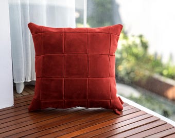Puede incluir: Un cojín decorativo cuadrado, de color rojo ladrillo, con un patrón de cuadrícula. El cojín está hecho de un material suave y aterciopelado y se coloca sobre una superficie de madera. El cojín está cerca de una ventana con una cortina blanca.