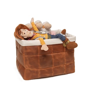 May include: A brown leather storage basket with a white lining. A toy cowboy doll with a yellow shirt and blue jeans is lying inside the basket. Two white plush sheep toys are also in the basket.